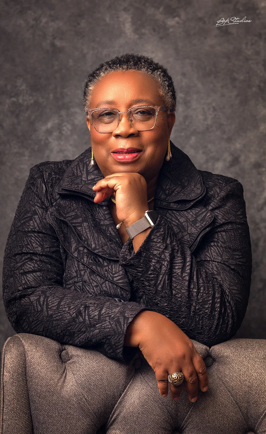 Folasade Adefisayo seated in a wing chair in a dark textured jacket, hand resting at her chin.